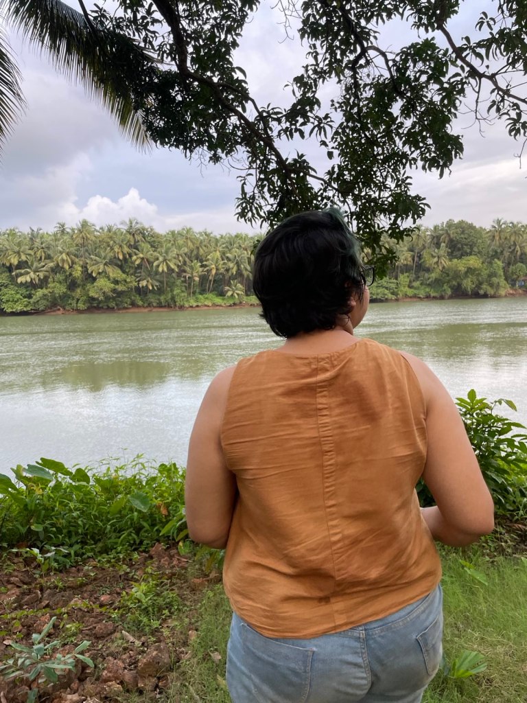 An image of Srinidhi standing at the bank of the river and looking away from the camera. She is dressed in a brown top and jeans. She has short cropped hair.